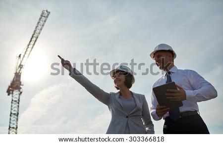 business, building, teamwork, technology and people concept - smiling man and woman in hardhats with tablet pc computer pointing finger up at construction site