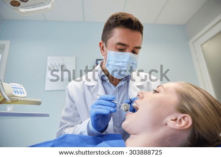 people, medicine, stomatology and health care concept - male dentist in mask with dental mirror and probe checking female patient teeth up at dental clinic office