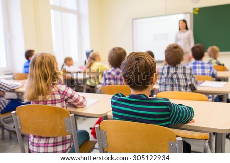 education, elementary school, learning and people concept - group of school kids sitting and listening to teacher in classroom from back