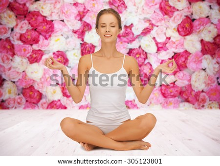 people, health, wellness and meditation concept - woman in underwear meditating in yoga lotus pose on wooden floor over wall of flowers background
