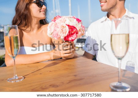 love, dating, people and holidays concept - smiling couple with bunch of flowers and champagne glasses and looking to each other at cafe