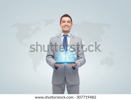 business, trade, people and technology concept - happy smiling businessman in suit holding tablet pc computer with hologram of shopping trolley over world map on gray background