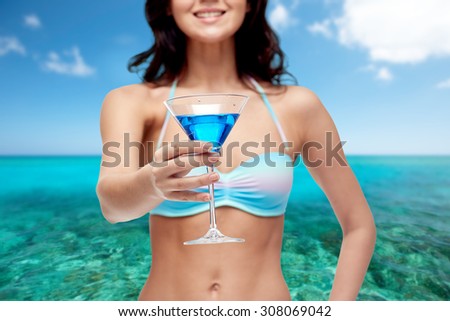 people, summer holidays, travel, tourism and alcohol drinks concept - happy young woman in bikini swimsuit holding glass of cocktail at beach party over sea and blue sky background