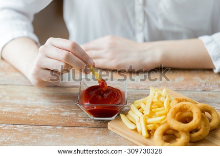 fast food, people and unhealthy eating concept - close up of hands with deep-fried squid rings, dipping french fries into ketchup bowl on wooden table