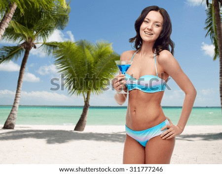 people, summer holidays, travel, tourism and alcohol drinks concept - happy young woman in bikini swimsuit holding glass of cocktail at party over tropical beach with palm trees background
