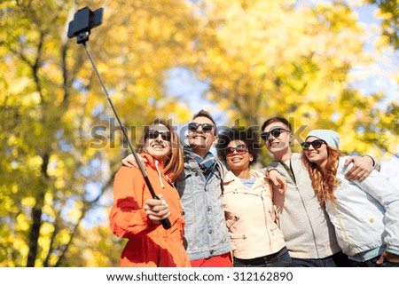 tourism, travel, people, season and technology concept - group of smiling teenage friends taking selfie with smartphone and monopod over autumn park background