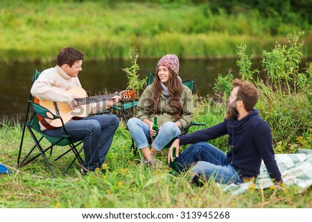 adventure, travel, tourism and people concept - group of smiling tourists playing guitar and drinking beer in camping