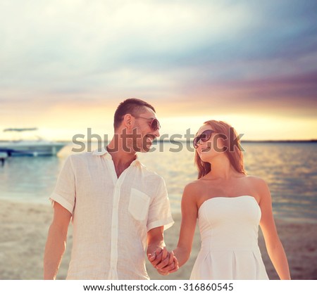 love, people, travel, summer and relations concept - smiling couple wearing sunglasses walking outdoors over beach background