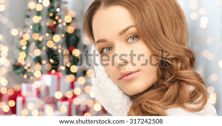 winter holidays, christmas and people concept - happy young woman in mittens touching her face over christmas tree lights background