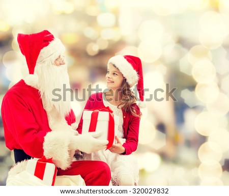 holidays, christmas, childhood and people concept - smiling little girl with santa claus and gifts over lights background