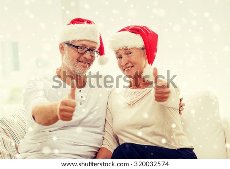 family, holidays, christmas, age and people concept - happy senior couple in santa helper hats sitting on sofa and showing thumbs up gesture at home