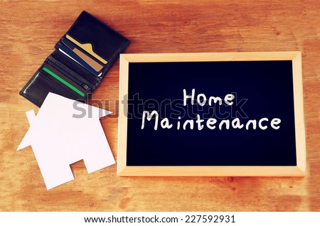 blackboard with the phrase home maintenance written on it over wooden board with wallet, credit cards and house shaped paper cut.