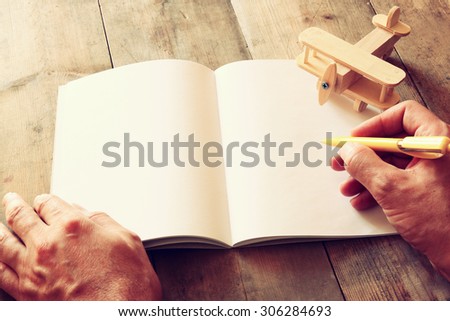 open blank notebook and man hands next to toy aeroplane on wooden table. retro style filtered image
