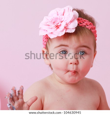 Caucasian brunette hair,blue eyed girl with a pink flower headband making a cute face, while eating her birthday cake  on a pink backdrop.