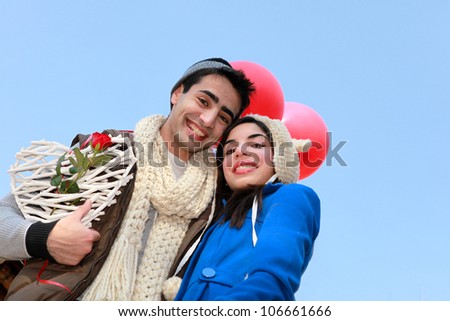 Attractive muslim couple or lovers smiling on Valentines Day date outside in the cold winter sky wearing beige brown and blue warm coats hats and scarves holding red balloons and heart with red rose