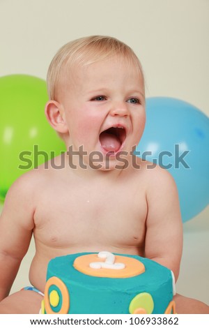 Blond hair blue eyed baby boy sitting by blue yellow and orange polka dot two tier birthday party cake yelling screaming and laughing in delight with blue green balloons behind on cream background
