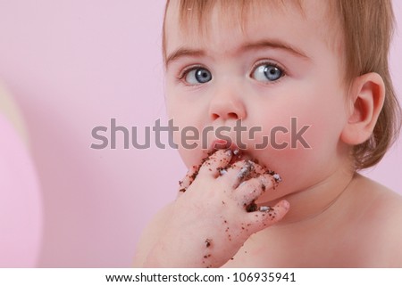 Cute happy brunette baby girl with blue eyes is tasting the chocolate cake and purple butter icing on her sticky fingers from her first birthday cake smash while sitting on a pink background sucking