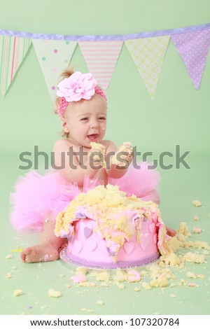 Adorable cute laughing blond hair baby girl with flower head band in pink tutu grabbing vanilla sponge cake with pink and purple heart icing while sitting on green background with flag bunting behind