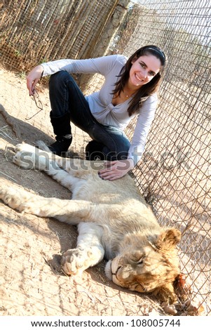 Young pretty brunette woman wearing grey and blue clothes sitting by beautiful wild lion cub stroking fur and smiling outside in nature park under blue sky
