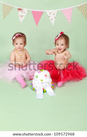 Two baby girl identical twin sisters sitting on green seamless background behind decorated birthday cake wearing red and pink tutus and bunting flags in the background about to break and smash cake