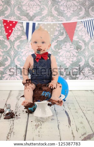 Adorable blonde haired boy with face covered in blue butter icing and sticky fingers eating cake at his birthday party