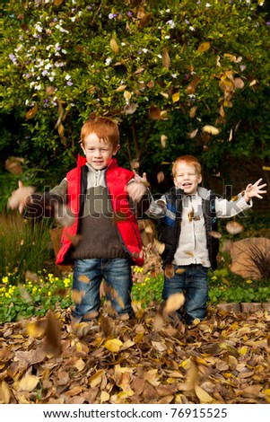 Two smiling brothers / sons are playing in the autumn / fall leaves, throwing them into the air and laughing in a park or garden setting