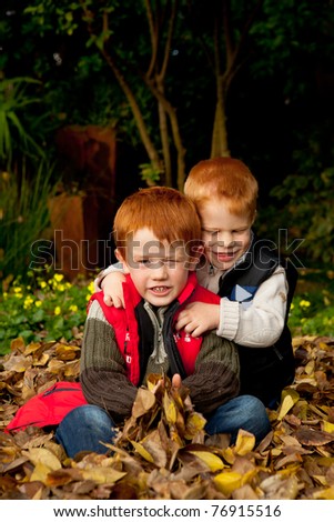 Two happy and smiling brothers or sons are sitting and hugging in a pile of colorful yellow and brown autumn / fall leaves in a garden or park setting