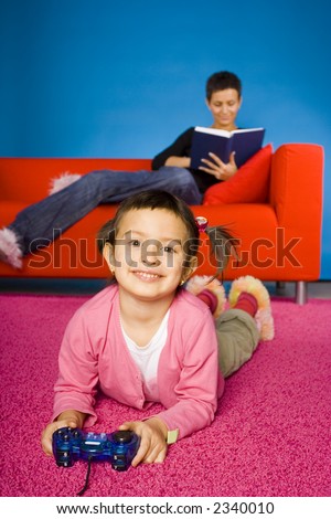 girl sitting on the carpet and playing video game and  her busy (with book) mother on the sofa