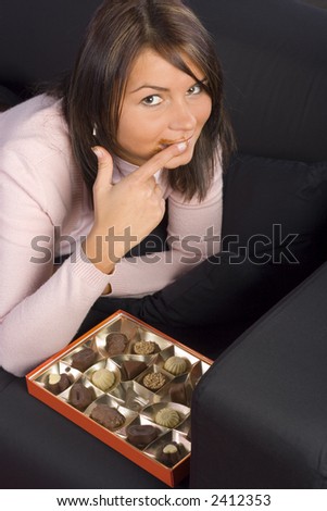 Young woman lying on the black sofa with box of chocolates; Licking her finger.