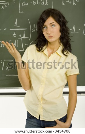 Teenage girl standing in classroom, in front of blackboard with hand up. Looking up