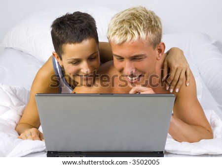Smiling couple lying in bed in front of laptop. Looking at monitor. Gray background, front view