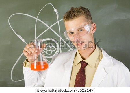 Young chemist in white apron. Holding beaker with orange liquid. Wearing goggles. Looking at camera, green background