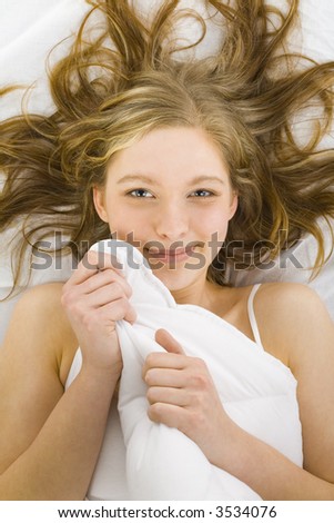 Young smiling woman lying in bed under duvet. Looking at camera, front view