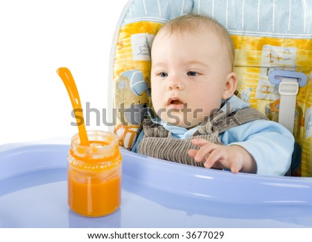 Pretty infant sitting on baby\'s chair. Looking at orange pulp in jar. White background