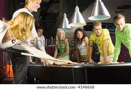 Large group of teenagers standing at pool table. Smiling and looking at right. One person is playing billard