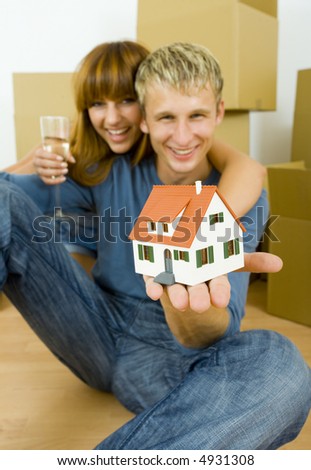 Young couple sitting on the floor in flat. They\'re looking happy. Woman is hugging man. They\'re looking at camera. man is holding house miniature. Focus on miniature. Front view