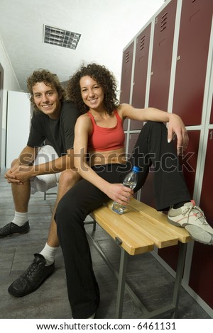 Young couple, sitting on bench in gym\'s locker room. Smiling and looking at camera. Woman is holding bottle of water