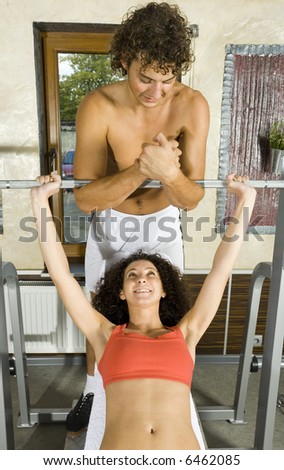 Young couple, working out in gym. Man is helping woman. Woman is lying on bench and picking up dumbbell. Smiling and looking at man. Front view