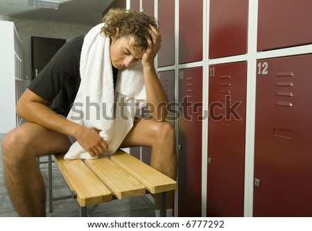 Young, exhousted man sitting on bench in locker room with closed eyes. Holding towel on neck. Front view