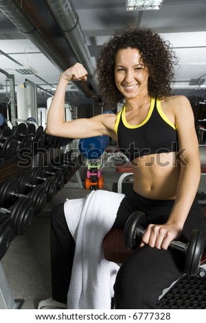 Young woman showing her\'s muscles. Sitting in gym with dumbbell in one hand. Smiling and looking at dumbbell. Front view