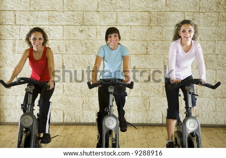 Three happy, young women working out on exercise bicycle at the gym. Front view, looking at camera.