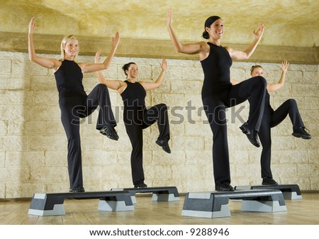 A group of women exercising in the fitness club. They\'re smiling and  have upraised hands and one leg. Low angle view.