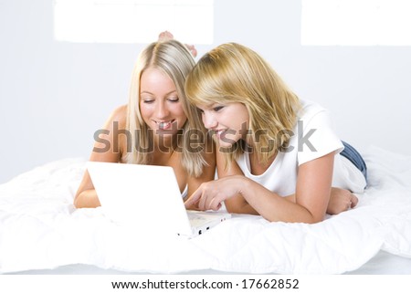Two girl friends lying in bed and do something on the laptop. Front view.