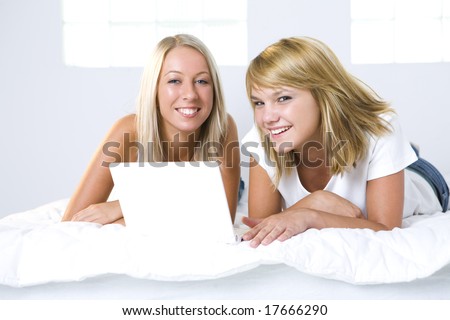 Two girl friends lying in bed and do something on the laptop. They're looking at camera. Front view.