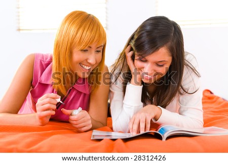 Two girls lying on bed and browsing magazine. One of them painting her nails. Front view.