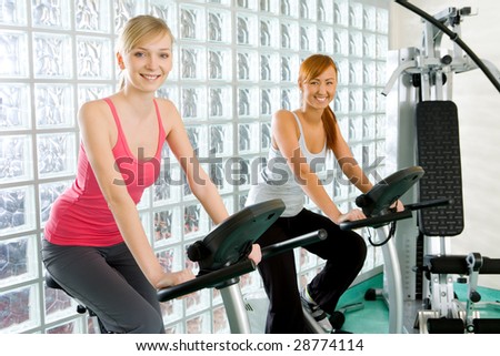 Happy women working out  on bike at gym. They're smiling and looking at camera.