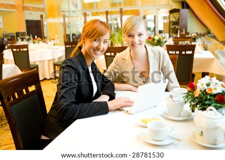 Two women sitting at table in restaurant. They\'re smiling and looking something on laptop.