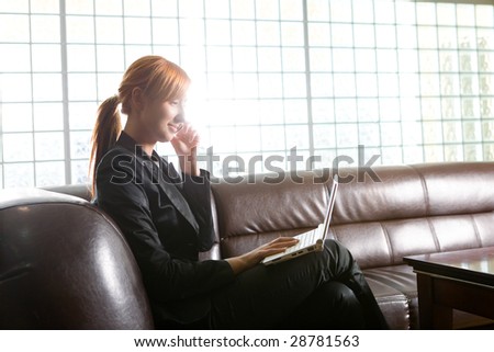 Woman doing something on laptop and speaking by cellphone. She\'s  sitting on couch.