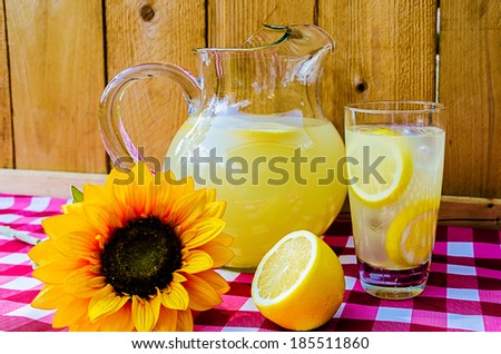 Lemonade with sliced lemons, pitcher, and sunflower on gingham table cloth.