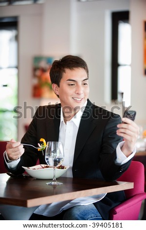 A young and attractive man uses his phone while eating a salad in an indoor restaurant